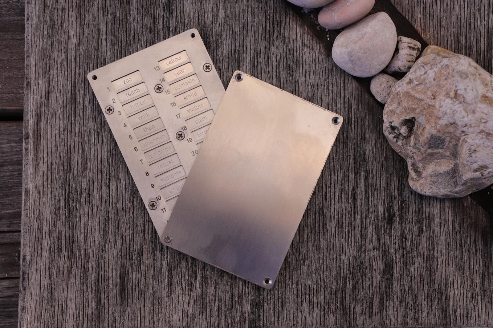 Geöffnetes SteelPhraze-Case aus gebürstetem Edelstahl auf verwittertem Holz: nummerierte, geätzte BIP39-Wörter unter der verschraubten Fixierplatte, die Deckplatte zur Seite gedreht, daneben Steine